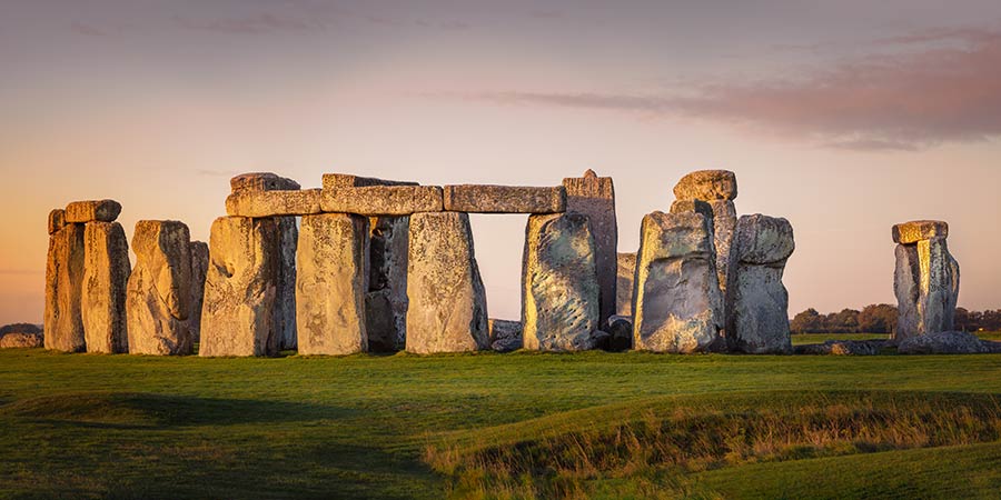 Image of Stonehenge, a circle of large, ancient stones standing in a grassy field. The stones are arranged in a pattern, with some leaning and others upright, with evening light. 